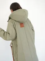 Abrigo largo color marrón con diseño acolchado de rombos, capucha y cierre frontal con botones a presión. Tiene bolsillos laterales y aberturas en los laterales inferiores.