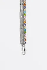 Collar de estilo gargantilla y largo, compuesto por múltiples cadenas plateadas. Dos de las cadenas están adornadas con cristales de colores brillantes (rosa, naranja, amarillo, verde, azul, violeta). Las otras cadenas son de eslabones finos. El dije principal es un aro metálico plateado con un corazón calado en el centro, decorado con pequeños cristales de colores.