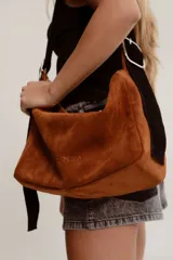 Bolso de hombro tipo hobo de gamuza color marrón óxido, con correa ancha ajustable de color negro y herrajes metálicos plateados. El nombre de la marca "GANESHA" está grabado sutilmente en el cuerpo del bolso.