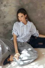 Camisa de corte recto y relajado, con cuello clásico, mangas cortas con detalle de ajuste y cierre frontal con botones. Presenta un diseño de rayas verticales en tonos celestes, blancos y grises.