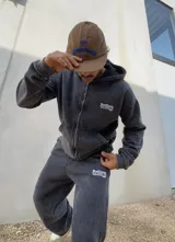 Conjunto de dos piezas compuesto por un buzo con capucha y cierre frontal y un pantalón de jogging, ambos confeccionados en tejido con efecto acid wash en color gris oscuro. El buzo cuenta con bolsillos frontales y logo bordado, mientras que el pantalón presenta un corte holgado y logo bordado en la pierna.