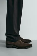 Bota estilo cowboy de caña media, confeccionada en piel con acabado de gamuza en color marrón oscuro. Presenta horma en punta, cierre lateral y suela con tacón cuadrado bajo.