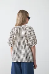Camisa de viscosa con estampado de lunares pequeños en negro sobre fondo blanco o crema. Presenta escote en V con abertura y botones frontales, y mangas cortas abullonadas.