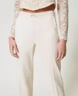 Pantalón de vestir color crema, de corte recto y tiro alto, confeccionado en tejido de punto milano con mezcla de viscosa elástica. Presenta bolsillos laterales inclinados, raya permanente y tira en la cintura con hebilla metálica con logo de la marca. Cierre lateral con cremallera.