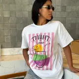 Remera blanca de manga corta con cuello redondo y un estampado frontal que incluye franjas verticales rosadas, un dibujo lineal de un cóctel con rodajas de cítricos y el texto "TANAT de Verano".