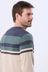 Remera de manga larga con cuello redondo, diseño a rayas horizontales en beige, marrón y azul marino, con finas líneas decorativas en rojo, azul y verde. Incluye un pequeño logo bordado en el pecho.