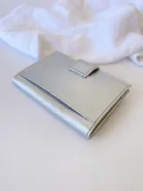 Tarjetero plateado de cuero con broche a presión. Tiene espacio para 4 tarjetas y billetes, y un bolsillo externo.