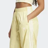 Pantalón deportivo amarillo pálido de corte holgado con cintura y puños elásticos. Presenta tres tiras blancas verticales a lo largo de cada pierna y logo de Adidas bordado en blanco en la cadera izquierda.