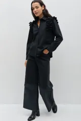 Camisa negra de manga larga con cuello clásico y detalle de volados en los hombros. Presenta cierre frontal con botones y puños abotonados.