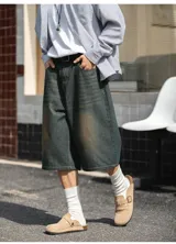 Bermuda de jean estilo baggy, tiro alto con cintura parcialmente elástica y cierre de botón. El lavado es oscuro con un efecto de decoloración horizontal en tono ocre/marrón claro en la parte delantera de las piernas.