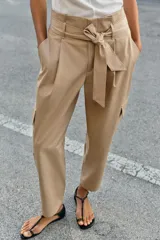 Pantalón de tiro alto estilo paperbag en color beige, con pinzas frontales y cinturón de la misma tela. Presenta bolsillos laterales tipo cargo con solapa y pernera ajustable con botones en el bajo.