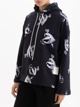 Buzo con capucha de corte holgado, confeccionado en tela de punto color negro con estampado integral de corazones atravesados por flechas en tonos grises y blancos. Presenta cordones ajustables en la capucha y mangas largas.