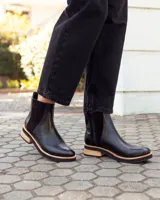 Botinetas tipo Chelsea de cuero negro, con paneles laterales elásticos y detalle de textura grabada en el talón. Presentan una suela plana con plataforma de madera clara y base de goma negra.