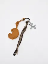 Llavero con diseño variado, incluye un dije de cuero sintético con forma de croissant color tostado, un cordón trenzado en tonos marrón y beige, y un dije metálico plateado en forma de perro globo.