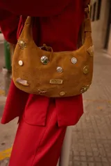Cartera baguette de gamuza negra adornada con diversos dijes y tachas metálicas en forma de sol, botones y animales, con correa de hombro y placa con logo metálico.