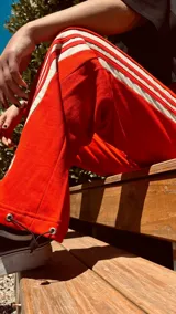 Pantalón deportivo color naranja con las icónicas tres rayas blancas laterales características de la marca Adidas.