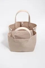 Bolso de mano grande tipo tote de Bimba y Lola, color beige, con doble asa superior y correa ajustable de tejido. Presenta logo metálico en la parte inferior y cierre superior.