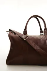 Bolso de viaje color marrón, confeccionado en cuero vacuno, con doble asa de mano y correa ajustable y desmontable. Cuenta con herrajes metálicos y costuras reforzadas.