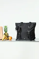 Bolso tipo shopper negro de diseño funcional, confeccionado en poliéster. Presenta correas reforzadas con detalles de argollas metálicas plateadas y un monedero pequeño removible enganchado en el frente. Cuenta con cierre principal y un diseño minimalista y versátil.