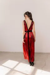Vestido largo rojo brillante con escote en V profundo, espalda abierta baja y breteles anchos. Presenta detalles en terciopelo negro que enmarcan el escote, recorren el frente y la espalda, y una faja doble en la cintura ajustable con cinto y hebillas. La falda tiene un corte frontal que permite ver sutilmente la pierna.