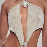 Top halter color beige claro, con escote profundo en V que se anuda en la parte inferior. Presenta un dije metálico con forma de espiral y centro nacarado en el centro del escote.