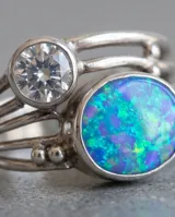 Anillo de plata 925 con diseño multicapa, decorado con una piedra central de ópalo sintético en tonos azules y verdes, y una piedra redonda translúcida más pequeña en la parte superior.