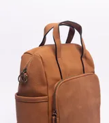 Mochila de diseño versátil en color marrón, confeccionada con una combinación de texturas en cuero sintético. Presenta un compartimento principal con cierre, un bolsillo frontal amplio con cierre y bolsillos laterales abiertos. Incluye manijas superiores fijas y una correa larga extraíble y ajustable para usar como bolso o mochila.