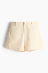 Short de tiro alto en denim de algodón levemente elástico, color beige, con bolsillos delanteros de parche con ribete y cierre frontal con botón.