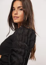 Blusa negra de tejido de punto acanalado, con cuello redondo y mangas largas abullonadas de tela semitransparente. Presenta una hilera vertical de pequeñas cuentas plateadas en el frente.