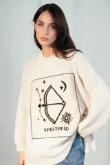 Sweater de tejido de punto crudo con diseño oversize y cuello redondo. Cuenta con un gran parche rectangular bordado en el frente con motivos astrológicos de Sagitario, incluyendo un arco y flecha, estrellas, la luna, el sol y la inscripción 'SAGITARIO'.