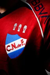 Camiseta de fútbol Umbro del Club Nacional de Football, modelo Away 1. Diseño principal en color rojo con cuello en V azul, detalles blancos en los hombros y puños blancos. Incluye el escudo del club, logo de Umbro y sponsors estampados.