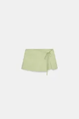 Falda pantalón de tiro medio en color verde claro, confeccionada con mezcla de lino. Cuenta con diseño cruzado en el delantero y detalle de lazada lateral.