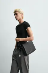 Bandolera de diseño rectangular en color negro con acabado texturizado. Cuenta con una solapa frontal con detalle metálico, cierre imantado y asa de hombro ajustable. El interior incluye un bolsillo plano y otro con cierre, además de un bolsillo plano adicional en la parte frontal.