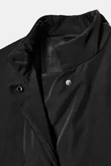 Chaqueta oversize de nylon en color negro, con cuello redondo subido, manga larga y cierre frontal con botones a presión ocultos. Presenta bolsillos delanteros y forro interior a tono.