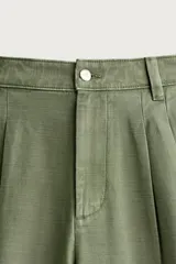 Pantalón chino baggy fit color verde militar, confeccionado en denim de algodón rígido, con corte holgado, tiro medio y pliegues frontales. Presenta bolsillos laterales y cierre frontal con cremallera y botón, con efecto lavado.