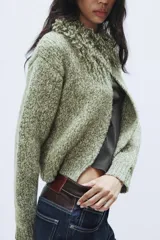 Chaqueta de punto de textura jaspeada en color verde, con cuello redondo adornado con flecos del mismo tejido, manga larga y cierre frontal con corchete metálico.