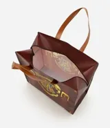 Bolsa tipo shopper de material plástico resistente, con diseño estampado que simula una maleta clásica con correas y hebillas. Presenta el escudo de Hogwarts en el centro y cuenta con asas dobles para llevar al hombro.