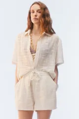 Camisa amplia y relajada de algodón liviano color blanco, de trama abierta y textura, con cuello camisero, mangas cortas y cierre de botones al frente.