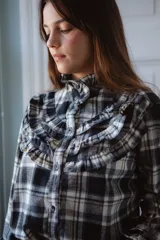 Camisa de manga larga con estampado de cuadros, cuello alto con volados y detalle de volados en el pecho. Presenta cierre frontal con botones oscuros.