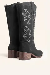 Bota de caña alta estilo tejano confeccionada en gamuza color marrón oscuro. Presenta detalles de costuras decorativas en forma de llama en la caña, taco cuadrado de madera y punta almendrada.