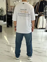 Remera oversize color beige, con mangas cortas y cuello redondo. Presenta un estampado tipográfico en la espalda con la frase "There are no facts, only interpretations" y otros detalles de diseño.