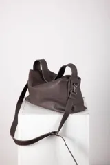 Cartera de cuero color marrón, con diseño suave y flexible. Cuenta con cierre metálico superior, doble asa corta para llevar al hombro y una correa larga regulable y desmontable. Interior forrado con bolsillos organizadores.