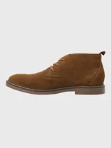 Bota tipo chukka de Hush Puppies, confeccionada en descarne color marrón, con cordones al tono, suela de goma y detalle de tirador en el talón con franjas rojas y negras.