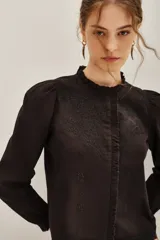 Camisa de algodón color negro con cuello alto tipo mao y volados frontales. Presenta bordados detallados en la parte superior del pecho y mangas largas con puños elásticos.