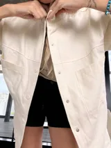 Sobre camisa color crema con capucha, cierre frontal con botones a presión, bolsillos con solapa en el pecho y mangas largas con puños abotonados.