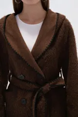 Chaqueta blanca de corte holgado con capucha, cierre frontal cruzado con botones y cinturón de tela ajustable. Presenta detalles de costuras decorativas en color negro en los bordes, solapas, bolsillos y cinturón.