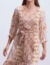 Vestido midi de gasa con estampado geométrico y floral en tonos naranja y turquesa sobre base beige. Presenta escote en V, cierre frontal con botones, mangas 3/4 con puños fruncidos y cintura elastizada con lazo de ajuste. El diseño incluye un ruedo con volado y corte asimétrico más largo en la parte posterior.