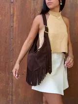 Cartera de hombro tipo hobo confeccionada en cuero gamuzado color chocolate, con detalle de flecos largos en la parte inferior y correa ajustable con hebilla metálica.