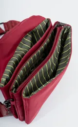 Bandolera de diseño rectangular en color verde oliva, con solapa frontal y logo de la marca grabado. Incluye una correa larga ajustable con el nombre de la marca tejido y un charm decorativo con forma de corazón y letra R en color rojo.