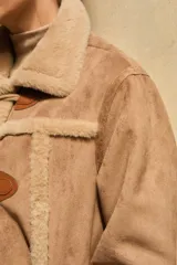Abrigo de estilo duffle coat en simil gamuza color tostado, con interior y bordes de piel sintética al tono. Presenta cierre frontal mediante alamares de cuero sintético y madera, cuello solapa y mangas largas.